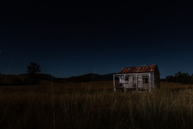 Abandoned shack standing in open field under starry sky. Perfect for themes of solitude, mystery, rural lifestyle, and night photography. Use in storytelling, background imagery, or mood setting for narratives depicting isolation or rustic environments.