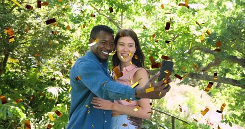 Joyful couple taking selfie with falling gold confetti in sunlit garden