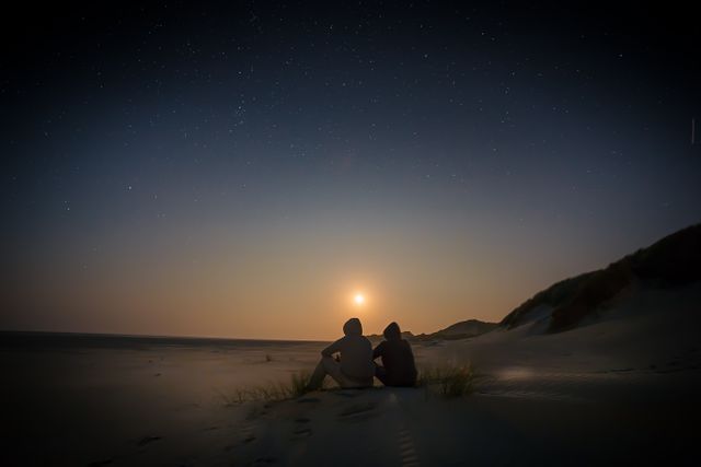 Silhouetted couple sitting on sand dunes as the sun sets and stars emerge in the evening sky. Ideal for use in themes about romance, nature, peaceful getaways, or outdoor adventures.