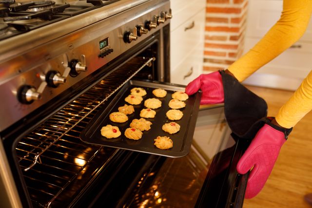 An african american woman baking cookies in the kitchen. she is taking a tray of cookies out of the oven while wearing pink mittens