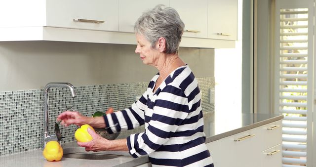 Senior woman washing vegetables in a modern kitchen, illustrating healthy living and home chores. Perfect for content promoting healthy lifestyles, home care products, senior well-being, and kitchen decor inspiration.