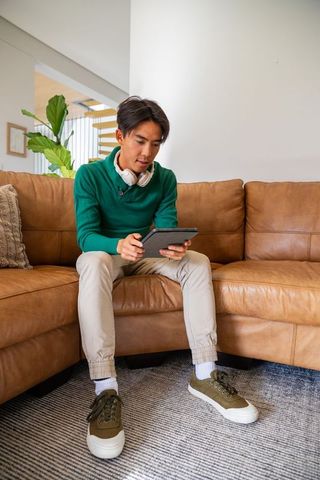 Asian man sitting on tan leather sofa using tablet with headphones in modern living room