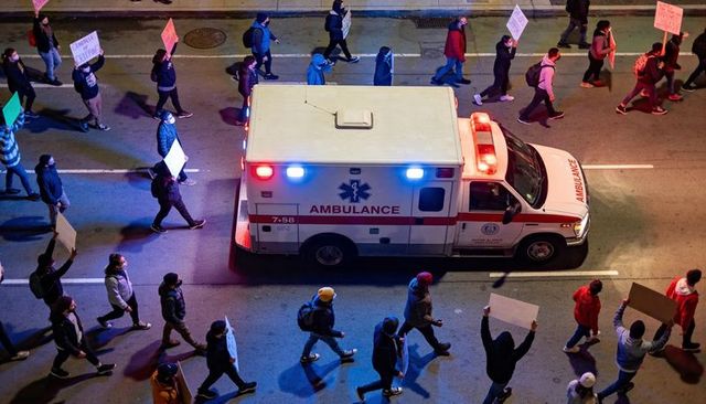 Ambulance navigating crowded protest march at night with flashing red and blue lights
