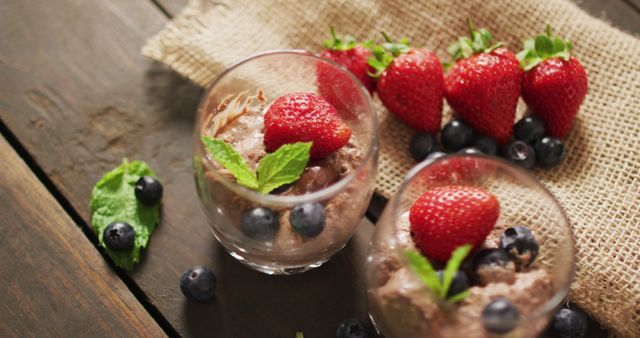 Appetizing chocolate mousse topped with fresh strawberries, blueberries, and mint leaves in glass cups on a wooden table. Ideal for use in food blogs, dessert recipe sites, and culinary magazines to highlight gourmet desserts and healthy eating options.