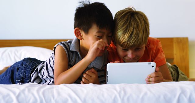 Two boys lying on bed and sharing a moment of laughter while looking at a tablet screen. Ideal for illustrating themes of childhood, technology use among kids, friendship, indoor leisure activities, and joyful moments in everyday life.