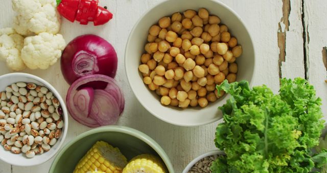 This image showcases a variety of healthy mixed vegetables and legumes on a rustic wooden table. The assortment includes chickpeas, leafy green lettuce, red onion, corn on the cob, white beans, and cauliflower. Ideal for use in nutritional articles, healthy eating blogs, recipes, and menus highlighting plant-based diets. Promotes natural and organic food choices.