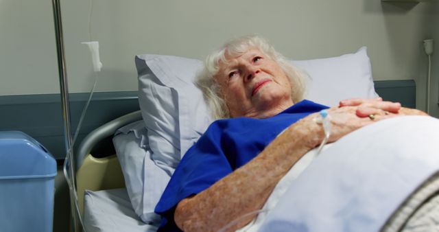 Elderly woman lying in hospital bed with IV drip attached, looking up thoughtfully. Potential use for healthcare services, rehabilitation centers, medical ads, and articles about elderly care.