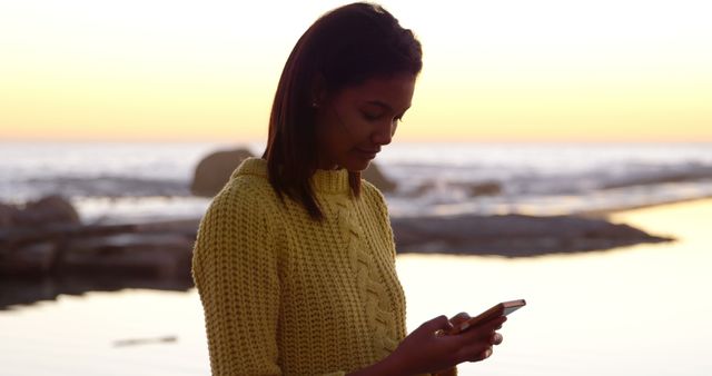 Image depicts young woman standing alone at the beach, texting on her phone during sunset. Can be used for themes related to technology use in natural settings, peaceful leisure time, solo outdoor activities, digital communication, and serene beach moments.