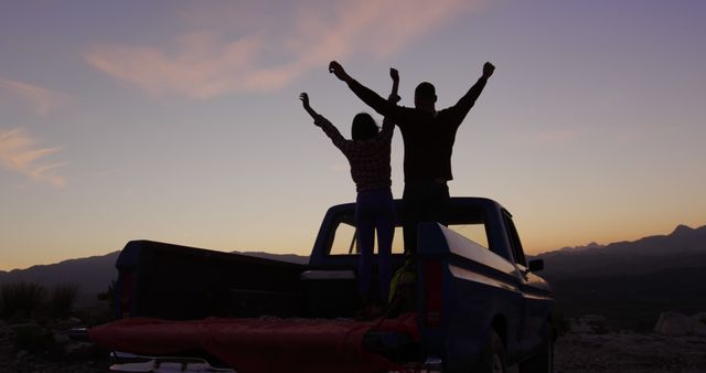 Couple standing in back of pickup truck raising arms in celebration. Beautiful mountain horizon and sunset sky in background. Perfect for themes related to adventure, travel, romance, outdoor activities, freedom and journey.
