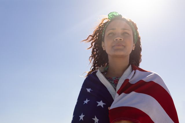 This image depicts a biracial woman wrapped in an American flag, looking forward with a determined expression. The clear blue sky and sunlight create a sense of optimism and hope. This image can be used for themes related to patriotism, national identity, freedom, and diversity in the USA. It is suitable for use in advertisements, social media posts, and articles celebrating American values and independence.