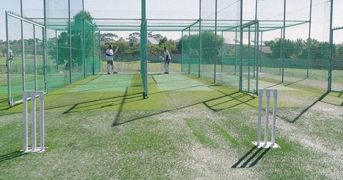 Two teammates practicing cricket in suburban net lanes with stumps, bats and protective gear