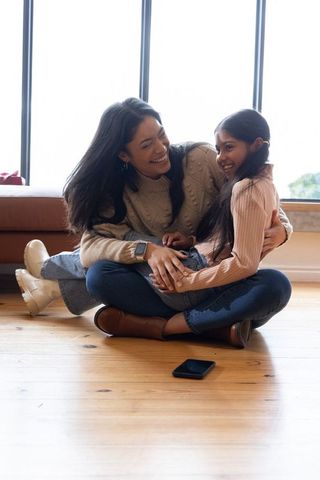 Laughing asian mother hugging school-age daughter on wooden floor with smartphone nearby