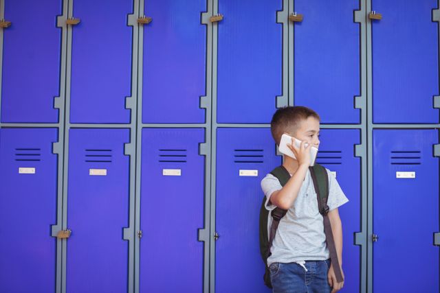 Elementary school boy standing in front of blue lockers, talking on a mobile phone. He is wearing casual clothing and a backpack, indicating a school setting. This image can be used for educational materials, technology in education, school communication, or childhood lifestyle themes.