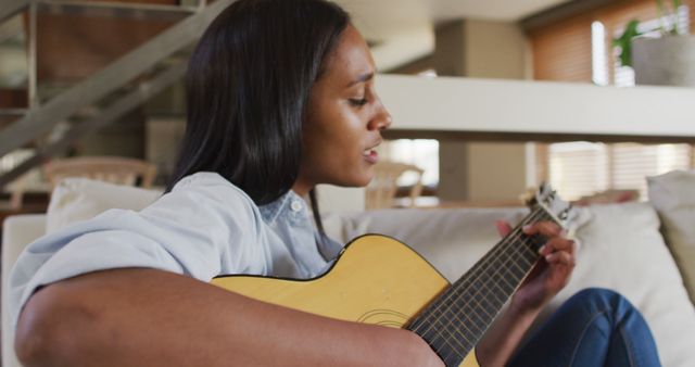 Young woman sitting on couch playing acoustic guitar. She is focusing and possibly singing. Home environment with a modern, cozy ambiance. Ideal for websites or articles about music, hobbies, home relaxation, and lifestyle. Could be used in ads for guitar lessons, music apps, or home furnishings.