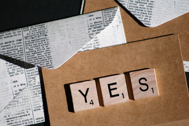 Scrabble tiles spelling ‘YES’ placed on Kraft paper background with newspaper envelopes. This image exudes a vintage and retro theme, apparent with the aged, handmade materials used. Ideal for use in articles, blogs, or social media posts focusing on positivity, acceptance, handmade gifts, DIY crafts, or vintage aesthetics.
