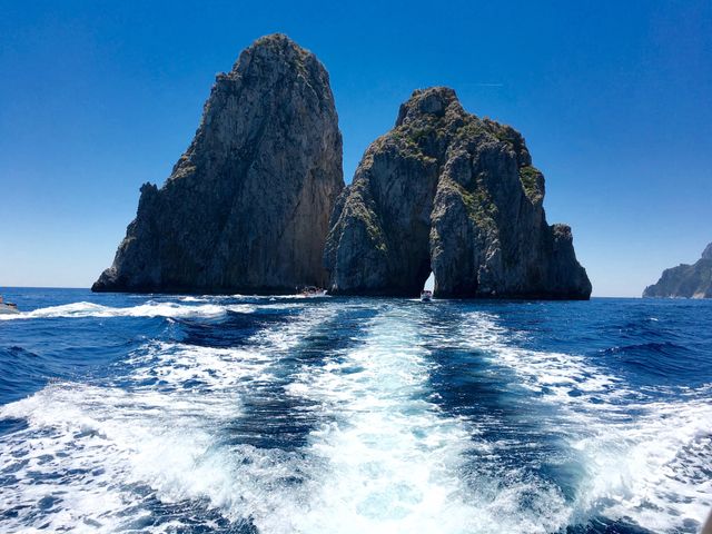 A picturesque view showcasing the famous Faraglioni rock formations rising from the Mediterranean Sea. The wake of a boat leads the viewer's eye toward the rock formations, adding dynamic movement to the scene. The clear blue sky and vivid waters make this image perfect for travel brochures, nature-inspired publications, or advertisements promoting tourism in Italy. Ideal for illustrating adventures or vacation experiences in stunning landscapes.