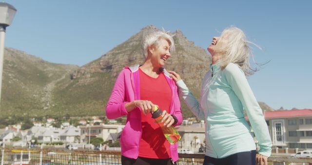Two senior women enjoying a moment of laughter during their morning exercise routine outdoors. They are dressed in activewear, holding a water bottle, with a scenic mountain and residential background. This image can be used for promoting healthy lifestyles, senior fitness programs, or the importance of friendship and social activities among elderly individuals.