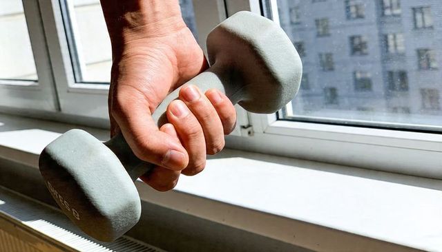 Close-up of hand holding gray neoprene dumbbell at sunlit windowsill in urban apartment