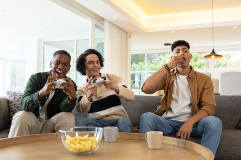 Three friends gaming and eating chips on couch with controllers in modern bright room