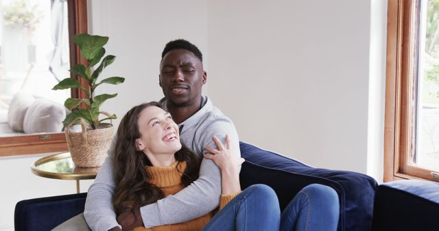 Interracial couple cuddling on sofa in modern living room. Ideal for promoting themes of love, diversity, and home living. Can be used in advertising for lifestyle brands, relationship blogs, or home decor.