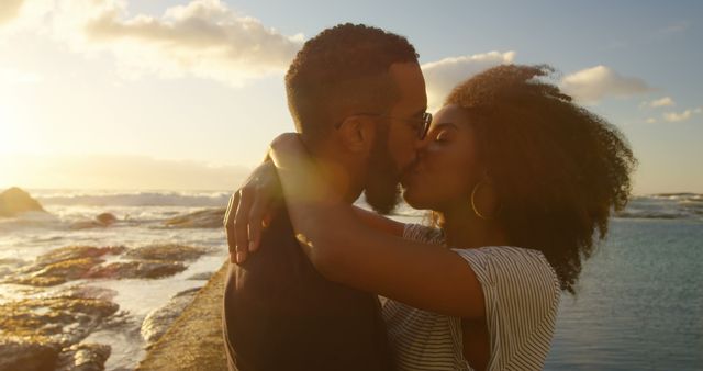 Romantic African American couple shares a kiss by the sea. Their affectionate moment captures the beauty of love in an outdoor setting.
