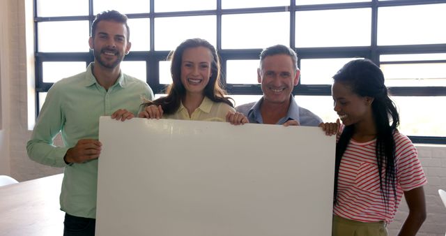 Diverse group of colleagues smiling and holding a blank whiteboard in a modern, bright office setting. Ideal for business presentations, team collaboration imagery, or showcasing a positive work environment.