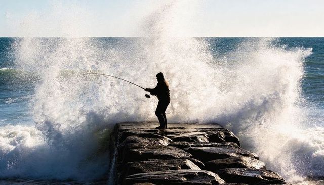 Silhouette angler casting from rocky jetty while ocean waves crashing into towering spray