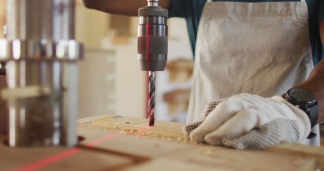 Image showing a craftsman using a drill press to cut into a piece of wood in a woodworking project. The person is wearing gloves for safety and focused on precision. Ideal for articles, tutorials, and promotional materials related to carpentry, DIY projects, woodworking tools, and safety practices.