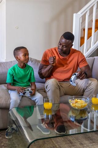 African American Father and Son Playing Video Games on Couch, Sharing Snacks and Smiles