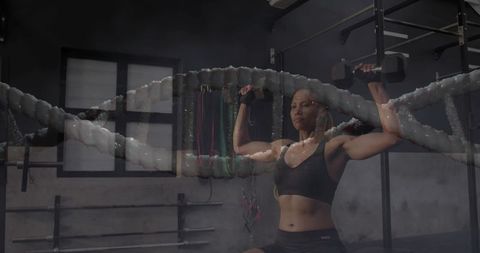 Female athlete pressing dumbbells overhead on bench in industrial gym with battle rope