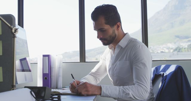 Businessman sitting at an office desk and writing in a notebook. Natural light streams in through large windows with a scenic mountain view. Ideal for workplace productivity illustrations, professional office contexts, or business-related projects.