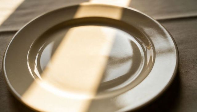 Glossy off-white ceramic plate catching warm diagonal sunbeam on brown textured placemat