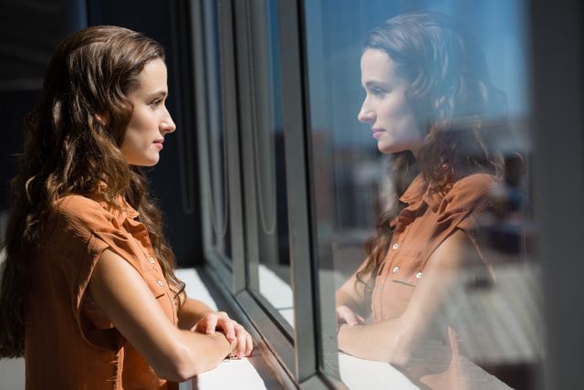 A businesswoman stands in an office, gazing thoughtfully through a window while her reflection is visible in the glass. This can be used in articles, blogs, and advertisements focusing on business, professional life, reflection, motivation, and workplace atmosphere.