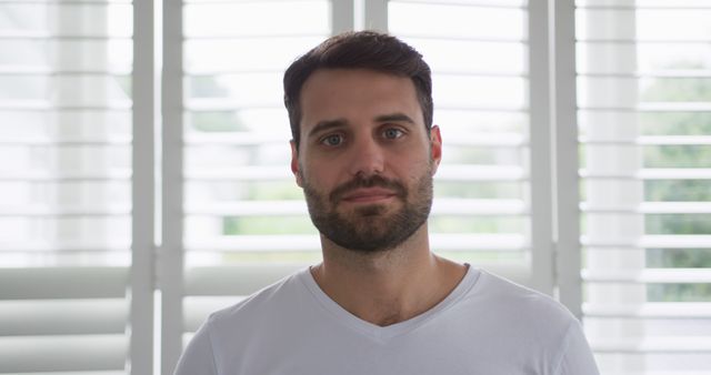 A man with a beard stands indoors wearing a white shirt, in front of window blinds. This image can be used for presentations or articles about home living, men's fashion, casual styles, or contemplative moments. It may also be suitable for professional portfolios or social media content.