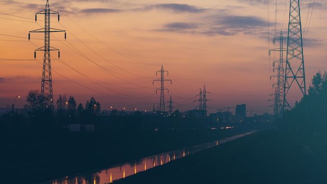 Beautiful scene featuring electric power transmission towers silhouetted against a vibrant sunset sky with a calm canal reflecting the light. Ideal for use in energy industry visual presentations, urban planning discussions, or environmental awareness campaigns.