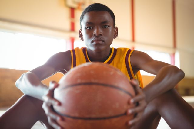 Teenage boy sitting on basketball court holding ball, showing focus and determination. Ideal for sports training, youth athletics, motivation, and basketball-related content.