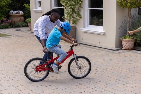 African american father teaching son to ride bike in suburban driveway with helmet