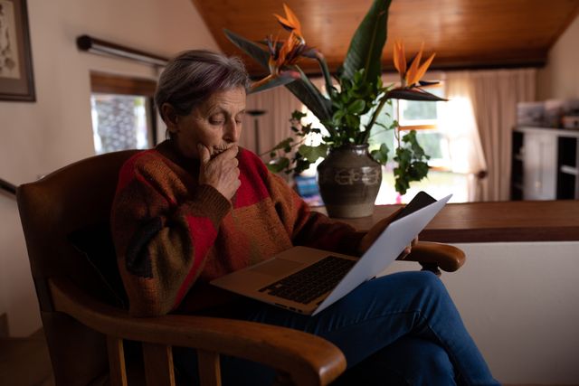 Side view of a senior Caucasian woman with short grey hair wearing a brown sweater sitting on an armchair in her sitting room, holding and using computer laptop, vase with flowers in the background.