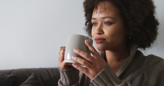 Happy biracial woman sitting on couch with mug and looking ahead in sunny living room. Lifestyle, wellbeing, relaxation, free time and domestic life.