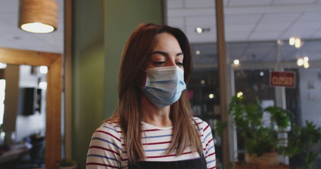 Woman wearing face mask working in coffee shop, emphasizing health and safety measures during pandemic. Suitable for topics about social distancing, hospitality industry, safety protocols, modern workplaces, and new normal.