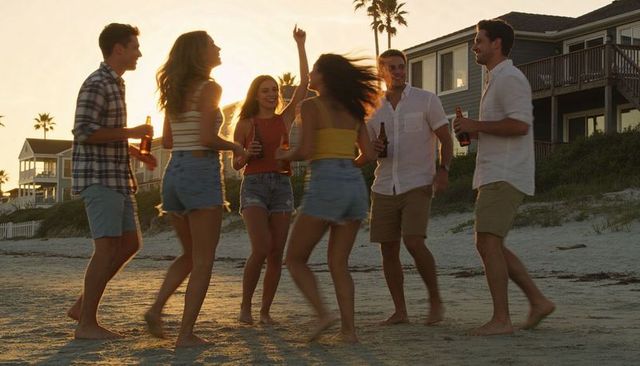 Friends partying on beach at sunset clinking beer bottles dancing barefoot golden hour