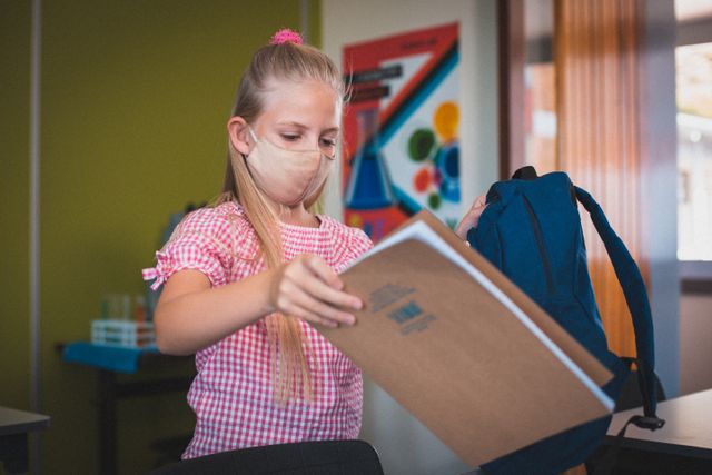 Caucasian schoolgirl wearing face mask holding book and bag in classroom. Ideal for educational materials, back-to-school campaigns, and content related to health and safety measures during the COVID-19 pandemic.