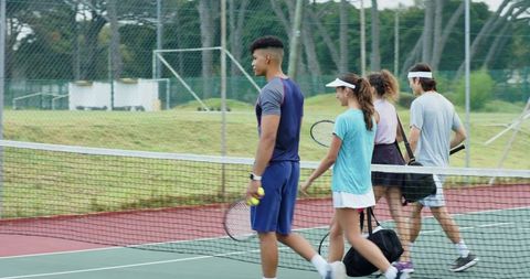 Young diverse tennis players walking across court carrying rackets, balls and sports bags