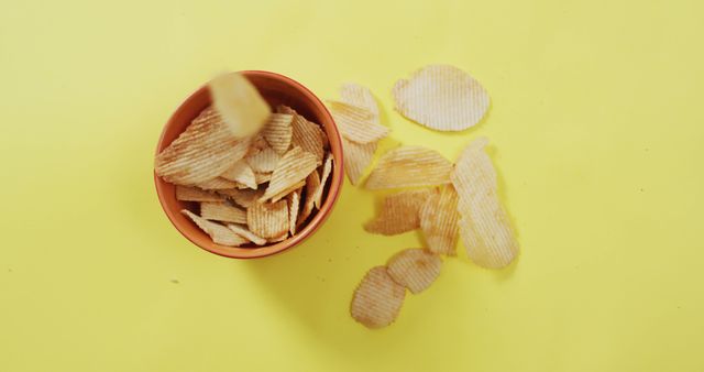 Potato chips, with ruffled texture, falling from a bowl on a yellow background. Ideal for illustrating snack foods, parties, junk food cravings, or casual eating moments. Use in advertisements, food blogs, social media posts, or health-related articles discussing occasional indulgences.