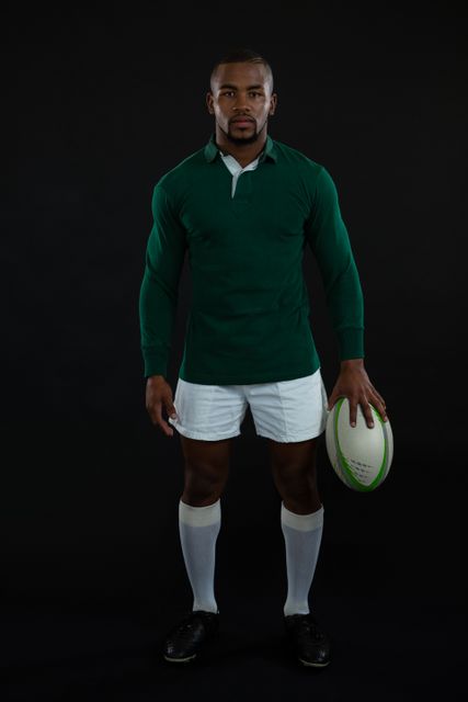 Male rugby player standing with ball against black background. Ideal for sports promotions, athletic apparel advertisements, fitness campaigns, and rugby-related content.