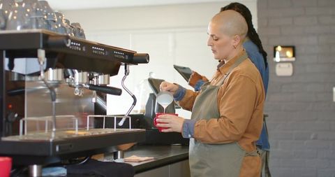 Barista pouring steamed milk into red cup behind espresso machine in urban cafe