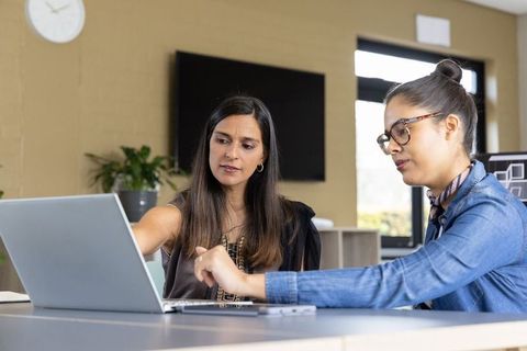 Diverse female coworkers collaborating on laptop in office mentoring teamwork productivity