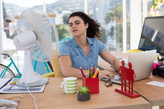 Female executive sitting at desk in modern office, enjoying breeze from table fan while working on laptop. Ideal for depicting workplace comfort, summer office scenes, productivity, and modern work environments.