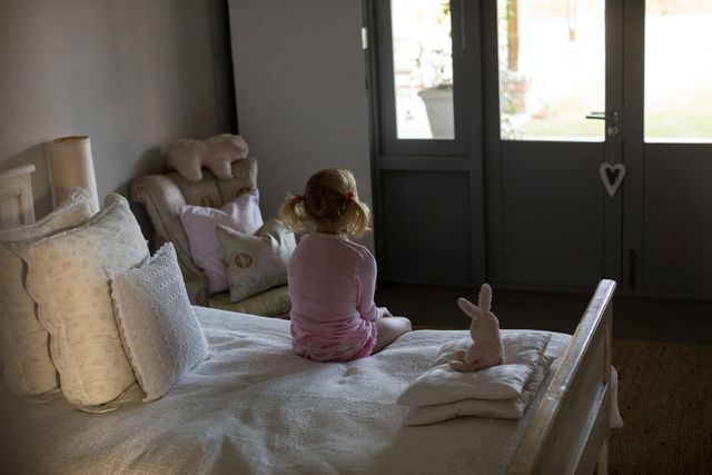 This image depicts a young girl sitting on a bed in a cozy bedroom, looking thoughtful. The room is softly lit by natural light coming through a window. Stuffed animals and pillows add a comforting touch to the scene. This image can be used for themes related to childhood, reflection, solitude, home life, and peaceful moments.
