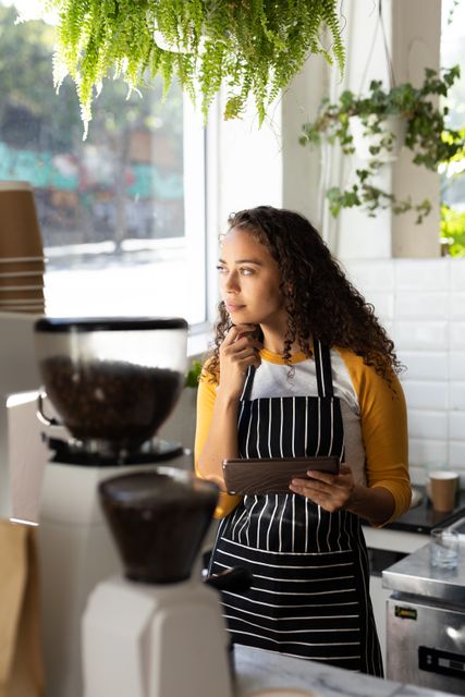 Thoughtful biracial female barista with hand on chin holding digital tablet looking away at cafe. unaltered, cafe culture, people, occupation and technology concept.